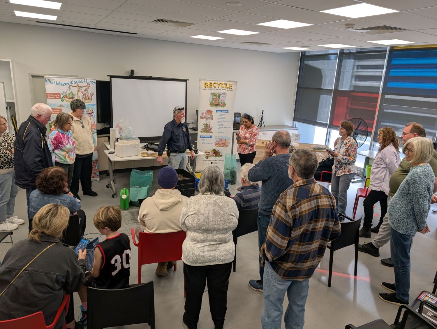 A diverse group of residents gathers inside a brightly lit classroom to watch a live worm farming demonstration led by an instructor